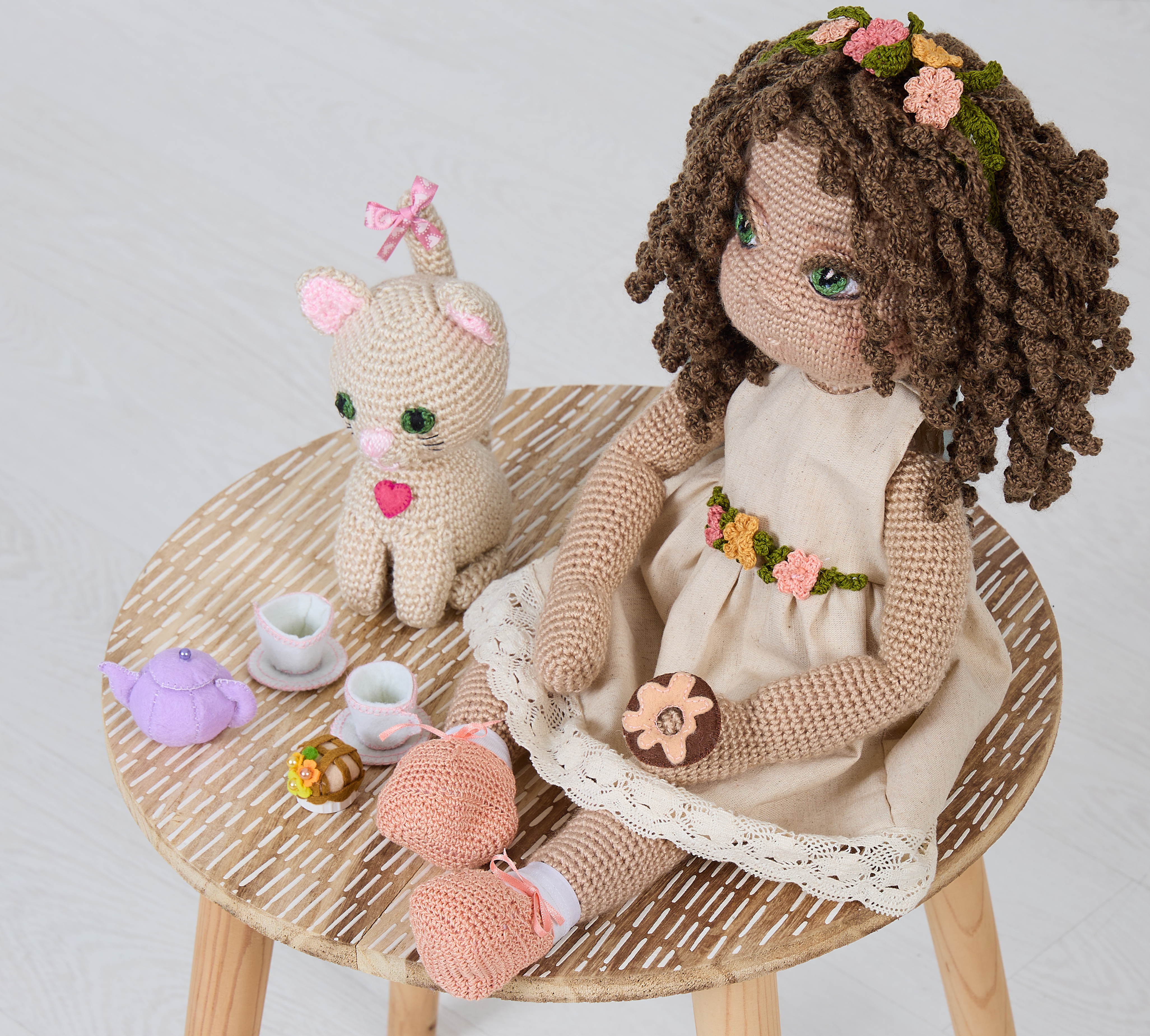 IMG_8102 (1) Nuestra muñeca artesanal de crochet, con cabello rizado castaño y una delicada corona de flores tejidas, viste un vestido color natural con detalles en encaje y un cinturón floral que realza su encanto. Sentada sobre un banco de madera tejido en espiga, comparte una encantadora merienda con una figura de gato amigurumi decorada con un corazón rosa y lazo en la cabeza. A su lado, una pequeña vajilla tejida en tonos pastel —tetera, tazas y dulces— completa este mundo imaginario lleno de calidez, juego simbólico y conexión emocional. Cada elemento está hecho a mano, pensado para estimular la imaginación y acompañar con dulzura momentos especiales. Propiedad de Mikadoll